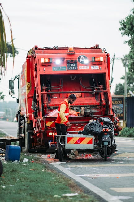 A worker in a yellow and red uniform operates a red waste collection truck parked at the side of a paved road in an outdoor setting during daytime. The truck's rear compartment is open, revealing mech