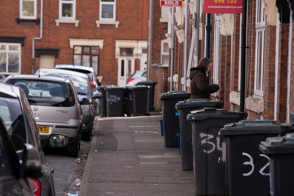 A middle-aged man with dark, slightly graying hair, dressed in a black t-shirt with white graphic text and black trousers, is seen disposing of waste into a large, silver, cylindrical rubbish bin on a